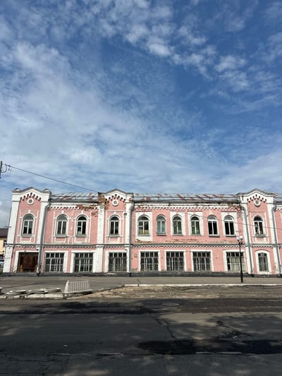 The image depicts a two-story, pink brick building with white decorative trim under a bright blue sky with scattered clouds. The building appears aged, with some sections of the facade showing signs of wear and damage, particularly along the roofline. It features numerous arched windows on the upper floor and rectangular windows with decorative metal grates on the lower floor. A dark wooden door is visible on the far left of the building's ground level. In front of the building, there is a paved road with some debris and construction materials, including concrete barriers and manhole covers. A single streetlamp stands near the center of the building's frontage. The scene suggests an urban or semi-urban environment, possibly in a historical district, located in Nogornyi, Russia. There are no people or discernible activity in the image.