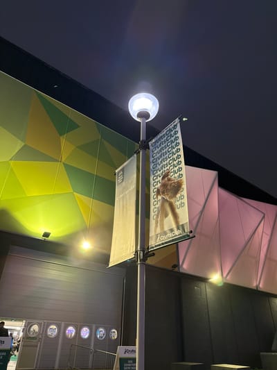 The image depicts an illuminated outdoor scene at night, located at the National Exhibition Centre (NEC) in the Metropolitan Borough of Solihull, United Kingdom.
A prominent grey lamppost stands centrally, topped with a spherical white light fixture emitting a bright glow. Attached to the lamppost are two vertical banners. The left banner is light beige and displays "Crufts Club" and "At your service." The right banner is white, featuring a large image of a light-brown, fluffy dog, with "Crufts Club" repeated vertically in green lettering and the "CruftsClub" logo at its base.
To the left of the lamppost, a building facade presents a striking geometric pattern in shades of green and yellow-green, brightly illuminated by yellow-green spotlights. Below this facade, a large, light-grey rolling door is visible. To its left, an entrance area with a smaller door and six circular windows shows signs of interior lighting. A green A-frame sign, partially visible at the bottom left, reads "Crufts DOG EXHIBITORS ENTRY DOOR." The head of one individual is partially visible near this entrance.
To the right of the lamppost, another building structure features a geometric facade illuminated with pink-purple lighting. The sky above is dark, indicating nighttime. The ground appears to be a dark, unidentifiable surface, with some low, dark rectangular structures visible on the far right. No explicit actions or interactions are discernible from the visible individual.