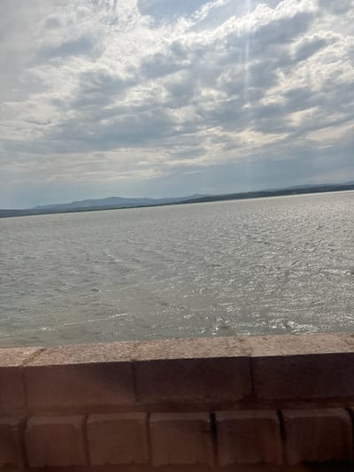 The image depicts a wide expanse of water under a cloudy sky. In the foreground, there is a textured brown surface, possibly a wall or embankment made of bricks. The water stretches to the horizon, where a low-lying landmass with hills is visible under a hazy sky. The sun's rays are breaking through the clouds, creating bright streaks of light on the water's surface. The overall scene is one of a natural landscape, likely a large body of water such as a lake or the sea, near a shoreline. It is highly improbable this scene is in the city of Port-la-Nouvelle, France, as the visible landscape does not match the known geographical features of that location, which is a coastal town in the south of France with a distinct harbor and beachfront.