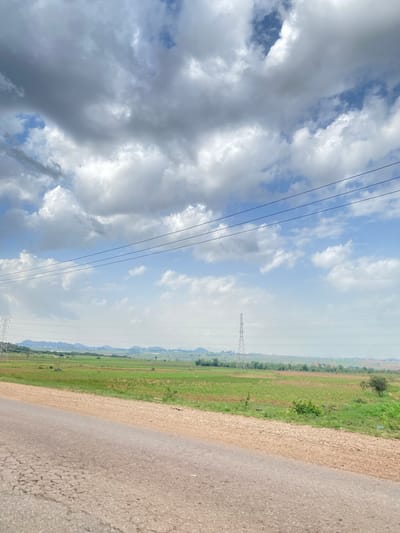 The image depicts a rural landscape in Barakin Tenti, Nigeria, under a sky filled with dramatic clouds. The scene is split diagonally, with the left side dominated by a vast expanse of sky and the right side by a road and a grassy field.
The sky is a mix of dark, stormy clouds and patches of bright blue, suggesting a dynamic weather pattern, possibly a transition between rain and sunshine. Several power lines stretch across the sky, indicating the presence of infrastructure in the area. In the distance, a large electricity pylon stands tall against the horizon, further emphasizing the utility infrastructure.
To the right of the power lines, a neatly divided landscape unfolds. A strip of lighter, possibly uncultivated land runs parallel to the road, giving way to a vibrant green field, indicative of agriculture or natural vegetation. The field stretches towards a distant, hazy horizon, where low hills or mountains are faintly visible. The road itself appears to be asphalted, with visible cracks and texture, suggesting it's a well-traveled route.
The overall impression is one of a broad, open rural setting, possibly captured during the daytime, given the brightness of the sky. The cloudy sky adds a sense of mood and atmosphere to the otherwise serene landscape. There is no visible text in the image.