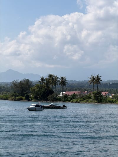 A tranquil coastal scene unfolds in Pemenang Timur, Indonesia, under a sky dotted with fluffy white clouds. The foreground is dominated by the calm, blue waters of the sea, with gentle ripples disturbing its surface. Two motorboats are anchored in the water. The closer boat is white with blue trim and a flag, while the second boat, positioned behind it, is darker with a similar design.
Beyond the water, a shoreline is visible, featuring a narrow strip of dark sand. Lush green vegetation lines the coast, with various trees and palm trees dotting the landscape. A cluster of buildings with reddish-brown roofs is situated amidst the greenery, suggesting a residential area or resort. In the background, rolling hills covered in dense foliage recede into the distance, with mist softening their outlines. The overall impression is one of peaceful, tropical serenity.