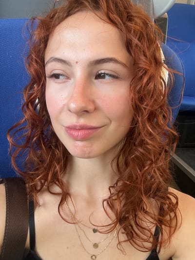 A close-up selfie shows a young woman with reddish-brown, curly hair flowing around her. She lies on her side, resting her head on a blue fabric surface, possibly a cushion or seat. Her head is tilted, and her gaze is directed towards the viewer, with a soft, subtle smile on her lips. Her skin is fair, with a healthy flush on her cheeks and a few small moles visible, particularly near her left eyebrow and cheek. Her eyes are light brown, and her eyelashes appear long, possibly enhanced with mascara. Her full lips are tinted with a natural pink shade.
She wears delicate gold necklaces with small charms, including a heart-shaped one, which are visible against her upper chest and collarbones. A black strap, possibly from a tank top or bra, is also visible at the bottom right of the frame. The background is mostly out of focus, dominated by the blue fabric directly beneath her and darker, indistinct shapes behind her head, suggesting an indoor setting. The overall mood is calm and pleasant. There is no visible text in the image. The location is contextualized as Stara Pazova Municipality, Serbia.