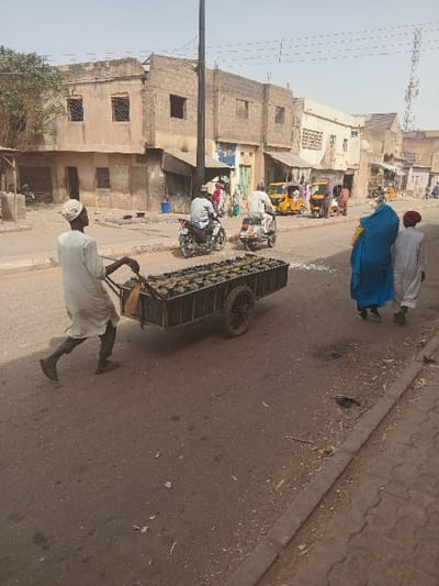 The image depicts a street scene in Dogon Nama, Nigeria, under a hazy, light-blue sky. In the immediate foreground on the left, a male individual, dressed in a light-colored long tunic and white skullcap, pushes a two-wheeled wooden handcart loaded with numerous dark, possibly rectangular items arranged in rows. He walks towards the right side of the frame. On the right foreground, two individuals walk along a dirt and concrete sidewalk. One wears a vibrant blue, full-body garment and appears to carry a yellow object on their back; the other, to their right, wears a light-colored tunic and a red cap. The paved road in the midground features two motorcycles with riders and two yellow tricycle taxis (keke napep), all moving or positioned away from the camera. The background consists of a row of two-story buildings constructed from concrete blocks or rendered earth, primarily light brown or beige, with multiple windows, some open and dark, others with grates. Utility poles and overhead wires are visible, and a lattice telecommunications tower stands in the distant right. The road surface and sidewalk show scattered debris and dirt.