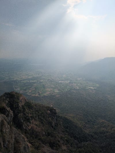 The image displays a panoramic view of a mountainous landscape, identified as overlooking the area of Kadaladi, India. The foreground features a steep, dark, rocky mountainside covered in dense, dark green and brown vegetation, primarily on the left and lower-center. This slope descends into a wide valley in the midground, characterized by a patchwork of light green agricultural fields and darker forested sections. Faint structures, possibly indicative of small settlements, are discernible within the valley. The background consists of distant, hazy mountain ranges. The sky occupies the upper portion of the image, appearing overcast with lighter clouds in the upper right. Prominent crepuscular rays, or sunbeams, are visible descending through the diffused light and atmospheric haze from the upper right quadrant. No human activity or specific man-made objects, aside from the cultivated land patterns and potential distant structures, are identifiable.