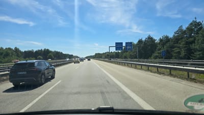 The image shows a view from a vehicle driving on a multi-lane highway. The sky is blue with some wispy clouds. On the right side of the highway, there is a metal guardrail and a dense line of evergreen trees. There are several blue highway signs visible. One sign indicates directions to "Pirmasens," "Landstuhl-West," and the route "42." Another sign indicates directions to "Trier," "Bingenfeld," "Kusel," and "Hahn" with an arrow pointing right for "Hahn" and route "62." Further down the highway, another sign indicates "U11" and "U36" with an upward arrow. In the foreground, the dashboard of the vehicle is partially visible, including the windshield wipers. Several cars are on the highway, including a black SUV in the left lane with the license plate "MA VM 278." The location is identified as Ramstein-Miesenbach, Germany.