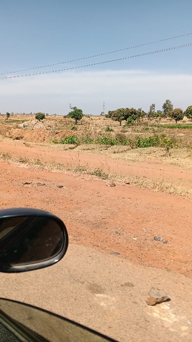 The image displays an open, arid, rural landscape situated in Barakin Kuru III, Nigeria, observed from the interior of a vehicle. The upper third of the frame is dominated by a clear, light blue sky traversed horizontally by two parallel strands of overhead power lines. A tall, metal lattice-work electricity pylon is visible in the distant midground, slightly to the left of center.

The midground and background consist primarily of reddish-brown earth with sparse, dry vegetation including patches of grass and small, leafy trees. Piles of reddish-brown soil are present, indicating ground disturbance.

In the foreground, a dark, oval-shaped side-view mirror of a vehicle occupies the lower-left quadrant, reflecting a blurred image. Below the mirror, the lower edge of a vehicle window is visible. The immediate foreground features a light grey-brown, possibly asphalt or compacted gravel surface, upon which a small, angular rock rests in the lower right. A reddish-brown dirt road or path diverges from this surface, extending into the midground. No individuals are present, and no active interactions or actions are discernible within the static scene.