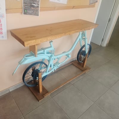The image shows a light blue bicycle frame that has been repurposed into a table. A wooden tabletop rests on the frame. The bicycle frame features two wheels, a suspension system, and handlebars, all painted in a consistent light blue. The wooden tabletop is unfinished and appears to be a live-edge slab. The entire structure is supported by a wooden base. In the background, a bulletin board with papers and a wall are visible. The floor is tiled with large, grey square tiles. The scene is indoors, likely in a public or commercial space. The location is identified as Barrio Estación, Spain, based on the provided context. There are no people visible in the image.