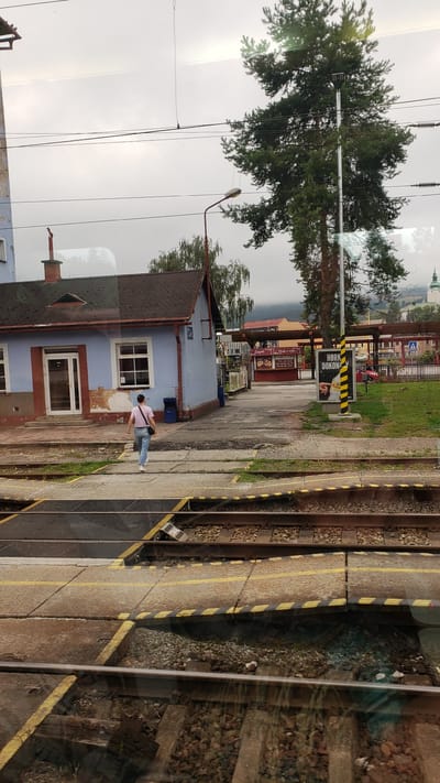 The image depicts a train station in Likavka, Slovakia. A woman wearing a pink t-shirt and blue jeans is walking away from a light blue building with a brown roof. She is carrying a black bag over her shoulder. In the foreground, there are railroad tracks with yellow and black striped crossing barriers. In the background, there are more buildings, trees, and a distant church steeple. A sign with "HORKO DOKONCE" is visible on a pole. The sky is overcast.