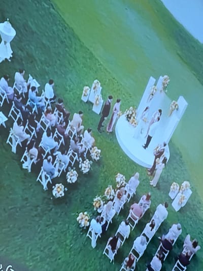 The image shows an outdoor wedding ceremony in what appears to be a grassy field. White folding chairs are arranged in rows facing a raised, circular platform. People are seated in the chairs, facing the platform where the wedding party stands. Bouquets of flowers are placed along the rows of chairs and around the platform. The location is implied to be near the city of Skalba Snt, Russia, indicated by the original request.