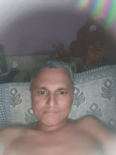 The image is a close-up selfie of a shirtless man with graying hair, lying down. He has a neutral expression and is looking directly at the camera. He is resting on a patterned fabric, possibly a bedsheet or quilt, with a floral design. In the background, there is a blurry view of a room, with hints of furniture and possibly decorations. A faint rainbow-like light flare is visible in the upper right corner. The overall setting appears to be indoors.
