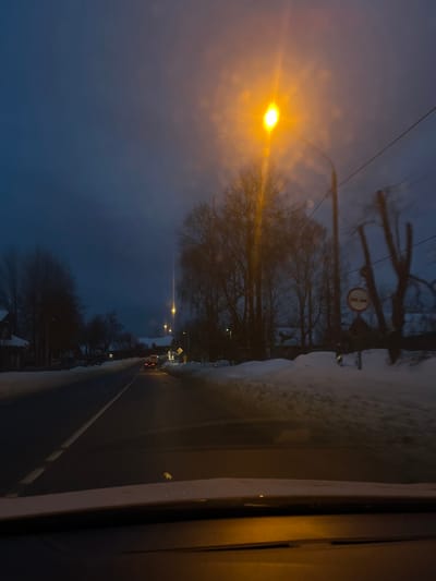 The image depicts a winter evening or early morning scene in Maksimkovo, Russia, viewed from inside a vehicle. The sky is dark blue-grey, with several street lights casting a yellow glow, most prominently a tall lamp on the right foreground creating a bright starburst effect. The asphalt road, appearing somewhat reflective, is marked with a solid white line on the right and a dashed white line indicating the center. Snow banks line both sides of the road, with a substantial accumulation on the right.
Ahead on the road, a vehicle with illuminated red taillights is visible, moving away from the viewer. Bare deciduous trees are prominent on both sides of the road, with denser groupings on the right. Scattered houses with snow-covered roofs are visible in the distance along the roadside. On the right, beneath the prominent street lamp and trees, a circular road sign with a red border and a horizontal white bar (likely a "No Entry" or similar prohibitory sign) is partially visible. Further along the right, a silhouette of a deer or elk-like figure is visible against a snow-covered surface. Power lines run horizontally across the upper right, attached to utility poles. The vehicle's dashboard and the base of the windshield are visible at the bottom of the frame.