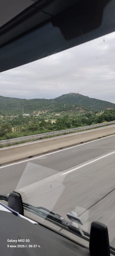 The image shows a view from inside a vehicle, looking out at a hilly landscape under an overcast sky. The foreground features the vehicle's dashboard, including a windshield wiper and part of the car's interior. The middle ground consists of a highway with concrete barriers and lanes. Beyond the highway, the landscape is covered with dense green vegetation, with scattered buildings and a communication tower visible on a distant hill. At the very top of the furthest hill, a structure resembling a fortification or ruins is discernible. The bottom left corner of the image displays text indicating "Galaxy M52 5G" and a date and time: "9 юни 2026 г. 06:37 ч." This date corresponds to June 9, 2026, at 6:37 AM. While the image itself does not explicitly show the city of La Jonquera, Spain, it depicts a roadside view consistent with the mountainous terrain surrounding such a location, likely captured during a journey.