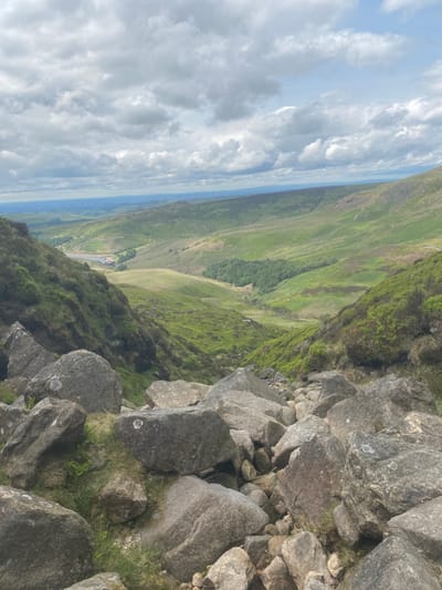 The image shows a view from the top of a rocky hill in Edale, United Kingdom. In the foreground, a collection of large rocks are scattered across the slope. The rocks are grey and brown with patches of green moss. The hillside is covered in green grass and vegetation. In the background, a valley with green fields and a small body of water is visible. The sky is partly cloudy with a few white clouds scattered across the blue canvas. The scene is peaceful and serene.