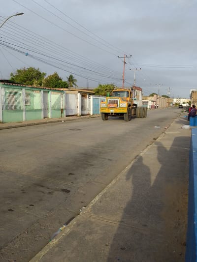 A yellow Mack truck with a red-painted structure is centered on a straight, concrete road in a neighborhood in Barcelona, Venezuela. The truck is headed toward the viewer. The street is lined with single-story homes behind white and green metal fences on the left, and a blue concrete curb on the right. Trash litters the side of the street. A couple sits on a concrete bench in the distance. Power lines cross above the street. Shadows of two people taking the photo are on the right side of the street. The weather is overcast.