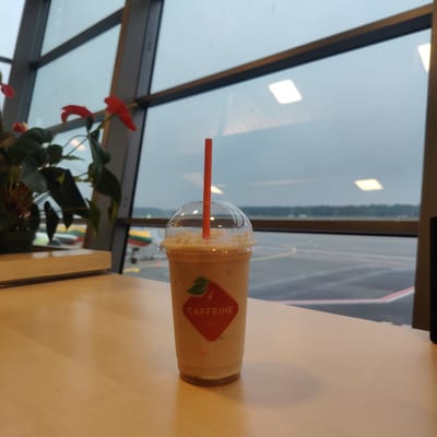An indoor scene in Skulte, Latvia, depicts a light-colored beverage in a clear plastic cup with a dome lid and an orange straw, resting on a light-colored wooden tabletop. The cup features a red and white logo reading "CAFFEINE EST. 2001," with a stylized leaf and lightning bolt. In the mid-ground to the left, a potted plant with dark green leaves and several red anthurium flowers is visible. The background is dominated by large, multi-pane windows looking out onto an airport tarmac under an overcast or twilight sky. Visible outside are yellow and red ground markings on the tarmac, and a portion of an airplane's fuselage or tail with yellow, green, and red stripes. Beyond the tarmac, a distant treeline forms the horizon. Reflections of interior square ceiling lights are present on the windowpanes.