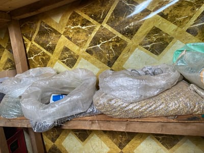 In the city of Theta ward, Kenya, on a wooden shelf, several large plastic sacks are stored. The sacks are transparent, allowing the contents to be partially visible. The sack on the right contains small, light-brown, granular material, possibly grains or seeds. To its left, another sack is visible, containing a dark, granular substance. Further to the left, two more transparent sacks are present, one appearing to hold a dark, granular material and the other a lighter material. Behind the sacks, a wall is decorated with a patterned wallpaper featuring a geometric design of diamonds in shades of brown and gold, with white marbling effects. The shelf itself is made of rough, unvarnished wood, and the backdrop suggests an indoor storage area.