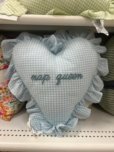 A light blue and white gingham heart-shaped pillow with a frilly ruffle trim is displayed. Embroidered in a teal-colored cursive font on the pillow is the phrase "nap queen." The pillow sits among other items including a green gingham item, a floral print fabric with an orange edge, and a tan corduroy object. Product information tags are visible on the items.