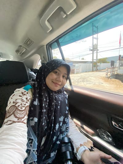 A woman wearing a black hijab and a white and brown patterned top is taking a selfie from the backseat of a car. She is smiling and looking at the camera. The interior of the car is visible, including the headrest of the seat in front of her, a car door, and a window looking out onto a street with buildings and utility poles. The scene appears to be in Mandar Sari, Indonesia, as indicated by the Indonesian flag visible in the background.