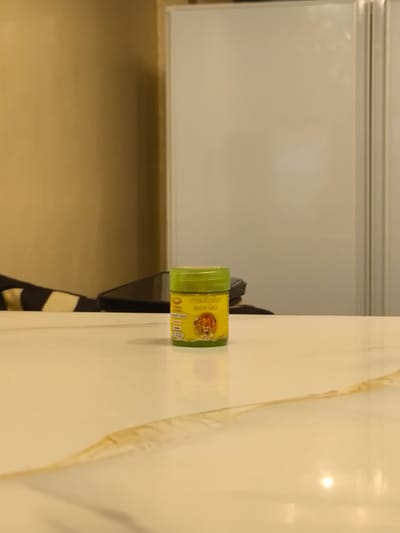 The image shows a small, green-lidded jar with a yellow label prominently displayed on a white, polished surface. The label features an image of a lion's head and text in what appears to be an Asian script. The jar is positioned towards the center right of the frame. In the background, a beige-colored wall meets a large, white, vertically oriented object with vertical lines, possibly a refrigerator or a cabinet. A dark object, potentially a phone or a laptop, is partially visible behind the jar. A portion of a striped fabric, black and yellow, is visible in the lower left corner. The lighting suggests an indoor setting with overhead illumination reflecting off the white surface. No people or specific landmarks in Imus, Philippines, are discernible.