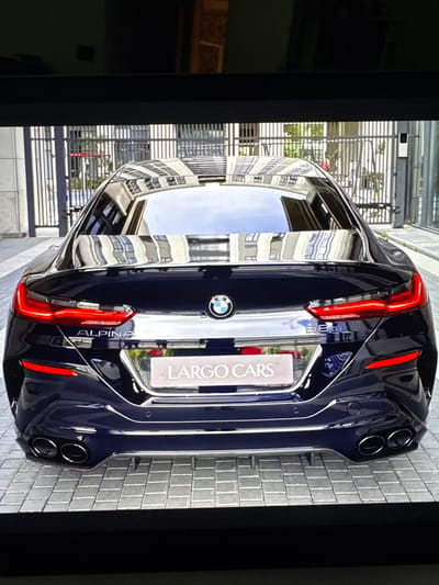 The image shows the rear of a dark blue Alpina B8 Gran Coupe parked on a paved surface. The car features distinctive red taillights, quad exhaust pipes, and badging that includes "ALPINA" and "B8 GT" on the trunk. A license plate frame reading "LARGO CARS" is also visible. The background suggests an urban environment with a building facade reflected in the car's windows and metal gates. The scene is devoid of people.