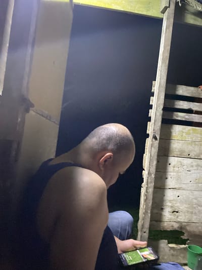 A man with a shaved head is sitting on the ground, looking at a smartphone. He is wearing a dark blue tank top and blue jeans. The background is dark, with some wooden structures visible. The scene appears to be outdoors at night. The location is identified as Lamtui, Indonesia, although this information is not visually verifiable from the image itself.