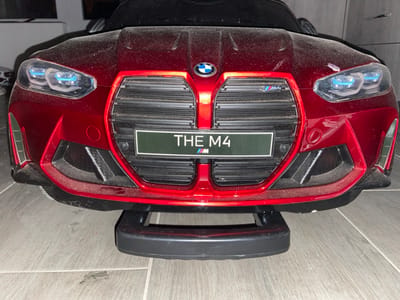 A dust-covered, metallic red toy vehicle, designed to resemble a BMW M4, is positioned facing forward on a light-colored tiled floor within the city of Breznitsa, Bulgaria. The vehicle's front features illuminated blue-tinted LED-style headlights and a large black kidney-shaped grille. A circular BMW emblem is affixed to the hood above the grille, and a smaller "M" logo is visible on the upper left portion of the grille. Below the central emblem, a dark green rectangular plate displays the text "THE M4". The entire visible surface of the toy car, including the red body panels, black grille, and lower bumper, is coated with a fine layer of dust. A black plastic support structure is present beneath the front bumper, and the background beyond the vehicle is dark and out of focus.
