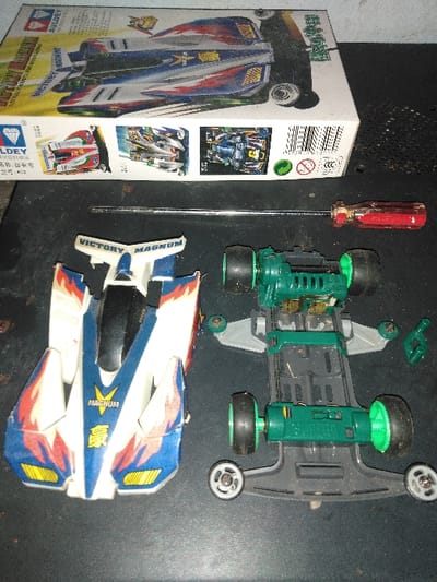 The scene, located in Mornington, Australia, displays an overhead view of mini 4WD model car components and accessories laid out on a dark, textured, possibly perforated surface. In the upper portion, a cardboard product box for an "AULDEY VICTORY MAGNUM" mini 4WD kit is visible, featuring an image of the assembled car, branding, Chinese text, and regulatory symbols including a barcode. Below the box, a metallic screwdriver with a red and clear plastic handle is positioned horizontally. To the lower left, the white and blue body shell of a "VICTORY MAGNUM" mini 4WD car, adorned with red and orange flame-like decals and Chinese characters, is presented in a disassembled state, showing signs of wear. To the lower right, the dark gray chassis of a mini 4WD car is visible, equipped with green plastic gear assemblies, four black rubber tires on green rims, and four small grey/white plastic guide rollers on the corners. A small, detached green plastic component rests near the chassis. No individuals are present in the frame. The components suggest a state of assembly, disassembly, or maintenance.