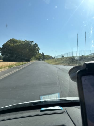 A clear, sunny sky is visible above a paved road. On the left side of the road, there is a field of dry grass and a large, dense tree. The road continues into the distance, where a white van is parked. On the right side of the road, there is a grassy embankment with a fence and some buildings in the background. The image appears to be taken from inside a vehicle, with a part of the dashboard and a phone mount visible in the foreground. There are no people visible in the image.
