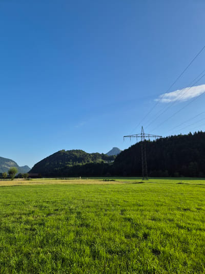 An outdoor, wide-angle photograph depicts a lush, green meadow in Niederndorf, Austria, under a clear blue sky with scattered wispy clouds. In the foreground, the meadow is densely covered in vibrant green grass, showing variations in height and texture. Beyond the meadow, a flat field extends towards a line of trees and rolling hills. A dark, silhouetted treeline forms the midground, with a prominent power line pylon and its accompanying wires cutting diagonally across the upper right quadrant of the image. The pylon is a tall, metallic structure with a lattice design. In the distance, a single, dark-peaked mountain is visible on the left side, partially obscured by haze. A building with a dark roof and lighter walls is situated behind a wooden fence in the midground on the left. The overall scene suggests a rural, natural environment with man-made infrastructure present.