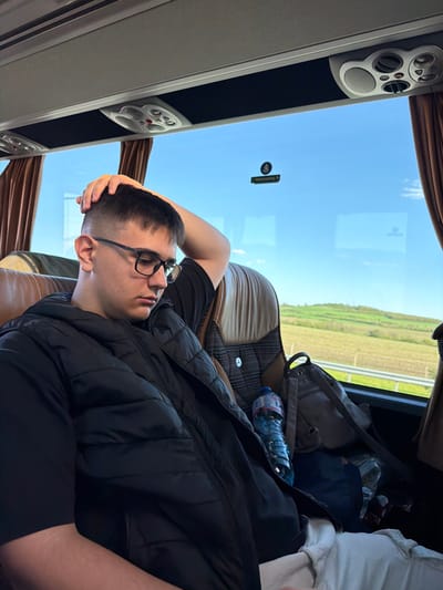 A young man with short dark hair and glasses is reclining in a bus seat. He is wearing a black t-shirt and a black vest. His right hand rests on his head. A water bottle and a grey backpack are visible on the seat beside him. The bus window shows a view of a green field and a highway. Overhead compartments and curtains are visible. A sticker with a symbol and the word "Nooduitgang" (Emergency Exit) are also seen on the window. The exact location in Yazdach, Bulgaria is not discernable from the image.