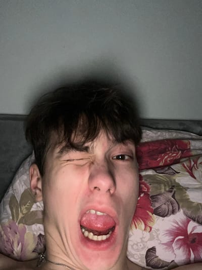 The scene depicts a close-up selfie of a young individual, likely male, located indoors in Zainsk, Russia. The subject's head occupies the majority of the frame, appearing to be lying down. Their left eye is tightly closed in a wink, while the right eye is open and directed towards the camera. The mouth is wide open, revealing the tongue and lower teeth, contributing to a contorted facial expression. The individual has short, dark, slightly messy hair. A thin, silver-colored chain necklace is visible around their neck, partially extending out of the lower frame. The subject is partially supported by a light-colored pillow on the left side of their head, and to the right, a pillow or duvet featuring a floral pattern with prominent red and brown/beige blossoms and leaves on a light background is visible. The background consists of a plain, light-colored wall, possibly light grey or muted green, displaying soft shadows behind the subject's head. The overall lighting is dim and even.