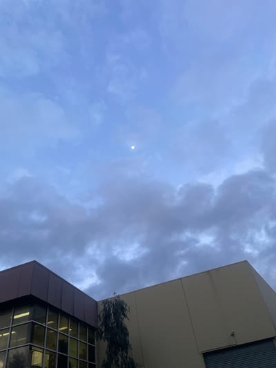 The image depicts a view of the sky and the upper portions of two industrial buildings in Northcote, Australia. The sky is a muted blue, filled with a mix of light and dark gray clouds. A small, pale crescent moon is visible in the upper right quadrant of the sky.

The building on the left has a dark brown, angular roofline and a facade with large glass windows. The windows reflect the dim light of the sky and appear to have blinds or shades covering them. Interior lights are visible through the windows, indicating activity within the building. A tall, thin tree with dark green foliage is positioned between the two buildings.

The building on the right is lighter in color, appearing beige or light tan. Its corner is also visible, extending towards the right edge of the frame. A grey roller door is visible at the bottom right of this building. A small, circular security camera is mounted on the exterior wall of this building.

The overall impression is one of a cloudy evening or early morning in an urban industrial area.