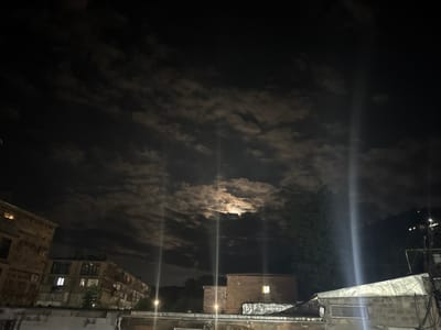 This is a nighttime photograph of the city of Kapan, Armenia, featuring a cloudy sky with the moon visible through a break in the clouds. The scene is dominated by the dark sky, with the moon casting a soft glow through the clouds. In the foreground, there are several buildings, some appearing to be apartment buildings with lit windows, suggesting human habitation. The architecture appears to be typical of older urban development. The overall atmosphere is nocturnal and somewhat atmospheric, with the moonlight creating dramatic lighting effects. There are no visible people or specific actions depicted in the image. The environment appears to be an urban setting, possibly residential or mixed-use, with some trees and vegetation also present. The photograph captures a moment of a cloudy night with the moon partially obscured, creating a visually interesting contrast between light and shadow.