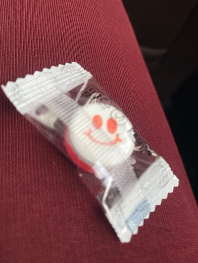 A single, individually packaged lollipop is depicted resting on a textured, burgundy fabric surface. The lollipop is round, primarily white with a red bottom edge, and features a printed smiling face with two large red oval eyes and a red curved mouth. It is encased in a transparent plastic wrapper with heat-sealed, serrated edges at both ends. Faint white text and graphics are visible on the wrapper. The background to the upper right is indistinct and dark, suggesting an indoor setting, potentially within a vehicle. No people are visible. The image contains no discernible visual information to identify the location as the city of Zarechnyi, Russia, or any specific geographical location.