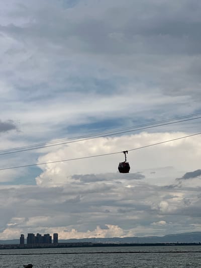 A lone red cable car hangs suspended from overhead wires against a cloudy sky. Below, a body of water, likely a lake or bay, reflects the overcast heavens. In the distance, a cityscape with numerous tall buildings lines the horizon, backed by a range of mountains. The scene is set in Xishan District, China.