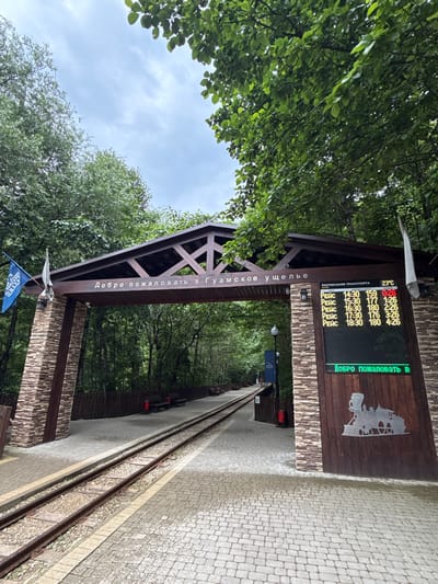 This is a photograph taken in Guamka, Russia, featuring a welcoming archway that marks the entrance to the Guam Gorge. The scene is set outdoors with lush green trees forming a canopy overhead, suggesting a natural, perhaps slightly humid environment. The sky is overcast, indicating a cloudy day.

The focal point of the image is a wooden structure that forms an archway. Above the arch, a digital display board is mounted, showing the schedule of transportation. The board indicates the current temperature as 23°C and lists departure times and numbers for several "REIS" (flights or trips). The text "Добро пожаловать в Гуамское ущелье" (Welcome to the Guam Gorge) is prominently displayed in Cyrillic on the archway and also on the digital board. A silhouette of a train is also visible on the wooden panel next to the digital screen, hinting at the mode of transport.

In the foreground, a paved pathway with cobblestone-like patterns leads alongside a set of train tracks. The tracks disappear into the distance, suggesting a journey into the gorge. A small blue flag with some text on it is planted near the base of the archway. The overall impression is one of arrival and anticipation, inviting visitors to explore the natural beauty of the Guam Gorge. There are no people clearly visible in the frame, focusing the attention on the entrance and the information provided. The image is oriented vertically, with the trees framing the top and right sides, and the archway and path extending downwards.