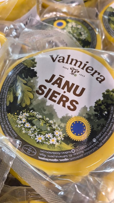 The image shows a close-up of a round cheese product, likely Jāņu siers, which is a traditional Latvian cheese often consumed during the summer solstice. The cheese is packaged in clear plastic, and a label is prominently displayed on the front. The label features the brand name "Valmiera" in large, dark brown lettering, followed by "JĀŅU SIERS" in even larger, bolder dark brown letters. Below "Valmiera" are the words "KOPS" and "1923", with a small illustration of a cow and some flowers.
The background of the label depicts a stylized forest scene with trees and a meadow dotted with white daisies. A circular emblem with the European Union stars and the text "GARANTĒTA TRADICIONĀLĀ RECEPTŪRA" (Guaranteed Traditional Recipe) is visible on the lower right side of the label.
Along the outer edge of the label, some nutritional information and text in Latvian are present. This includes "Ražotājs: AS Valmieras piens, Rīga", indicating the producer is Valmieras piens AS, located in Riga. Also visible is a code "LV P 9601 014 ES", which is a Protected Geographical Indication (PGI) or Protected Designation of Origin (PDO) registration number.
The image does not provide any information about the city of Līvāni, Latvia. The cheese is a packaged food product, and the scene is a still life of this product. There are no people or actions depicted. The environment is not discernable beyond what is shown on the product label.