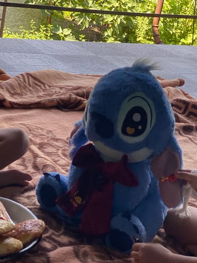 The image depicts a plush toy of Stitch from Disney's Lilo & Stitch, positioned on a brown blanket. The toy is blue with large eyes and is wearing a red bow. To the left of the toy, a person's legs are visible, and to the right, a child's hand is interacting with the toy, holding a bracelet made of colorful beads. A white bowl containing several baked goods, possibly pastries, is also present in the lower left corner. The background shows a tiled surface and green foliage, suggesting an outdoor setting. The scene is likely located in Lagodekhi, Georgia, given the provided location context.