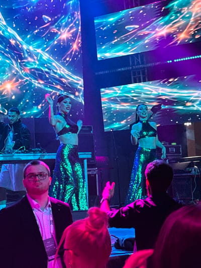 In Tsilkani, Georgia, a nighttime indoor event features a stage with vibrant, animated digital backdrops displaying abstract, nebulae-like patterns in electric blues, greens, and pinks. Two female performers in shimmering, sequined outfits, one in a green halter top and long skirt, and the other in a similar green top and skirt, are positioned center-stage, dancing with raised arms. To the left, a man in a black jacket operates DJ equipment. In the foreground, a man wearing glasses and a suit jacket with a "SECURITY" lanyard is visible, along with the backs of several attendees with brightly colored hair, suggesting a lively atmosphere. The lighting is predominantly blue and purple, contributing to the energetic and festive mood of the event.