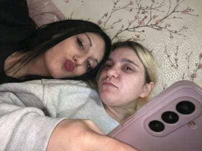 The image captures two adult females posing for a selfie indoors, located in the city of Lanchkhuti, Georgia. The female on the left has long dark hair and wears a black garment, exhibiting prominent eye makeup and red lipstick, with her lips puckered into a pout. She is resting on a light-colored surface, possibly a pillow. The female on the right has blonde hair and wears a light grey hooded sweatshirt. She is holding a pink mobile phone, which has three vertically aligned camera lenses on its rear, angled towards the viewer. Both individuals are looking towards the camera. The background consists of a wall adorned with light-colored wallpaper featuring a pattern of light pink floral branches, possibly cherry blossoms, against a subtly textured, light-toned base.