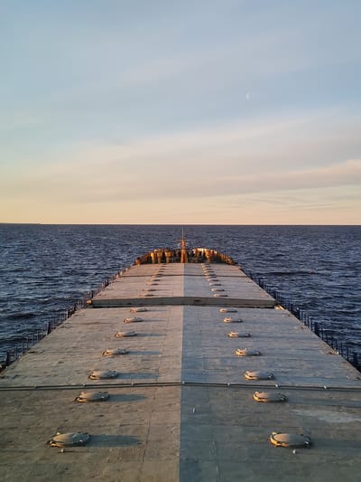 The image depicts a view from the deck of a ship or barge at sea. The deck is gray and features a symmetrical arrangement of circular hatches. A metal railing runs along each side of the deck. The sea stretches to the horizon, and the sky above is a gradient of light blue to pale yellow, with a crescent moon visible. The location is Myaksa, Russia. There are no people visible in the image. The focus is on the ship's deck and the seascape.
