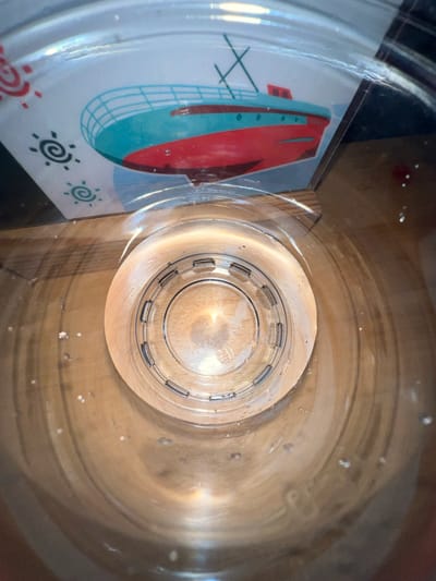 This media file is a top-down, fisheye lens perspective shot looking into a clear plastic container, possibly a water bottle or jar. Inside the container, a colorful graphic is visible on one side, featuring a stylized red and teal ship with a lattice structure and decorative sun-like symbols. The exterior of the container appears to be resting on a wooden surface, with the grain of the wood clearly visible and showing some wear. Small droplets of water or condensation are scattered on the inner walls of the container and on the wooden surface. The lighting suggests an indoor setting, with soft light illuminating the inside of the container, creating reflections and highlighting the texture of the wood. There is no discernible text in the image. The scene is static and focuses on the interior of the container and its immediate surroundings. The overall impression is one of a simple, everyday object, potentially in a domestic or storage setting. The location context provided is Ochamchire, Abkhazia.