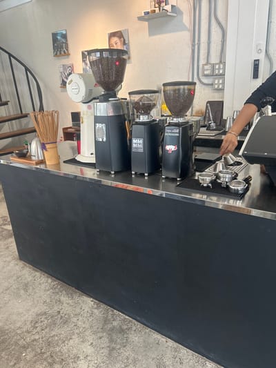 The image depicts a coffee shop counter in Khanom, Thailand. A person's arm and hand are visible on the right side of the frame, reaching towards a set of metal tampers and espresso portafilters arranged on a black mat. Behind the portafilters, three commercial coffee grinders are visible. The leftmost grinder is white and has a red and white logo. The other two grinders are black, with clear hoppers filled with coffee beans. The counter itself is black with a stainless steel top. On the left side of the counter, there is a metal kettle, a wooden board, and a bamboo whisk. In the background, there are shelves on the wall with framed pictures, and industrial-looking electrical conduits and switches. A spiral staircase is partially visible on the far left. The floor appears to be concrete.