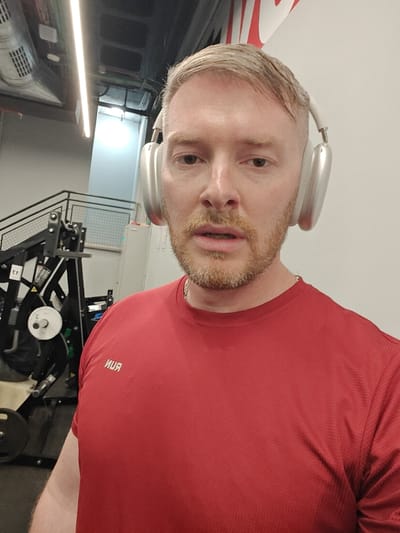 A male individual is depicted in a gym located in Nahariya, Israel. The man has light blonde hair, a short reddish-blonde beard, and a light complexion, appearing slightly flushed. He is wearing silver over-ear headphones and a red athletic t-shirt featuring the word "RUN" printed in reverse on the chest. A silver chain is visible around his neck. His mouth is slightly open. The background to the left shows black metal gym equipment, including weight plates and a pulley system, alongside a black metal staircase with a railing. Exposed ventilation ducts and fluorescent strip lighting are visible on the ceiling. To the right, a plain white wall is visible, with a partial red and white logo (resembling the Nike swoosh and lettering) near the upper right corner.