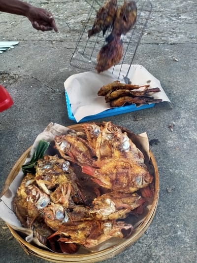 The scene is set outdoors in Celep, Indonesia, on a grey, cracked concrete or asphalt surface. A dark-skinned hand, visible from the wrist, holds a metal grilling rack containing several dark-brown, cooked fish, positioned above a light blue plastic tray. This tray is lined with beige parchment paper and holds approximately five smaller, golden-brown cooked fish. In the foreground, a large, round woven bamboo basket, also lined with beige parchment paper and garnished with green leaves, is filled with numerous larger, golden-brown fried or grilled fish, predominantly Tilapia, identifiable by their visible red fins and crispy texture. A portion of a red plastic container is visible on the far left. The hand appears to be engaged in the process of handling or transferring fish.