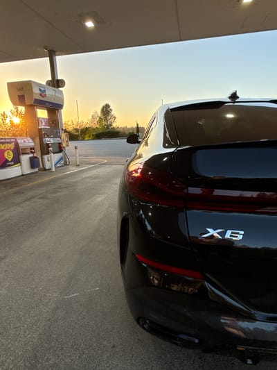 The image depicts a black BMW X6 parked at a Chevron gas station. The car is positioned on the right side of the frame, with its rear visible and the X6 emblem prominent. The gas station canopy and pumps are visible on the left, indicating the location is at a fuel service area. The sky suggests either sunrise or sunset, casting a warm light on the scene. Trees are visible in the background behind the gas station. It is possible that this is Parksville, Canada, but without additional information, it cannot be verified.