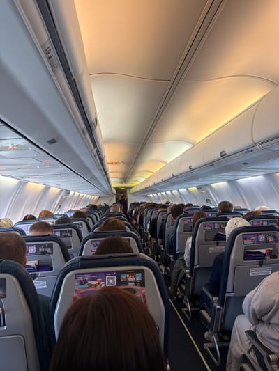 The image shows the interior of an airplane cabin filled with passengers. The overhead compartments are visible along the top left side of the image, with the row number "19" labeled on one. Passengers are seated in rows, their heads and the backs of their seats filling the frame. In-flight entertainment screens and advertising materials are visible on the seatbacks. The aisle stretches down the center of the image, disappearing into the distance. The emergency exit sign is visible further down the aisle. The photograph's location is not specified; there is no contextual information about the city of Arkhangel'sk, Russia.
