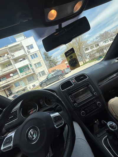 The image, taken from inside a car, presents a view of a street scene in Vetrino, Bulgaria. The car's steering wheel with the Volkswagen logo is prominent in the foreground, along with the dashboard controls, a VIP air freshener, and glimpses of a passenger in the vehicle. Through the windshield, several residential buildings with satellite dishes and air conditioning units are visible. A dark-colored car is parked on the street. The sky is partly cloudy, suggesting a sunny day. The in-car display shows the time 13:26 and mileage.