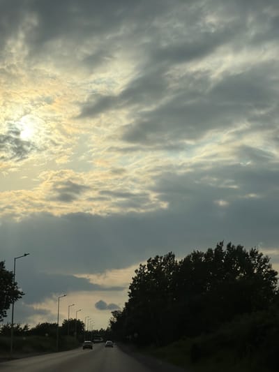 The image depicts a scene captured from inside a moving vehicle on a road in Mirikənd, Azerbaijan. The perspective is from the passenger seat, looking towards the right side of the road. The sky is filled with dramatic, textured clouds, with patches of sunlight breaking through, suggesting either sunrise or sunset. The weather appears to be overcast with some bright areas.

On the right side of the frame, a dark silhouette of trees lines the road, obscuring much of the landscape beyond. The road itself is visible, appearing wet, which might indicate recent rain or a humid atmosphere. Several streetlights are spaced along the side of the road, with the closest one prominently featured on the left side of the image. Further down the road, a few vehicles are visible, suggesting ongoing traffic. The overall mood is somewhat somber due to the dark clouds and silhouettes, yet the sunlight breaking through offers a sense of hope or transition. There is no visible text in the image. The time of day is likely early morning or late evening, given the lighting conditions. The presence of streetlights and the paved road indicate a developed area, likely a highway or main road.