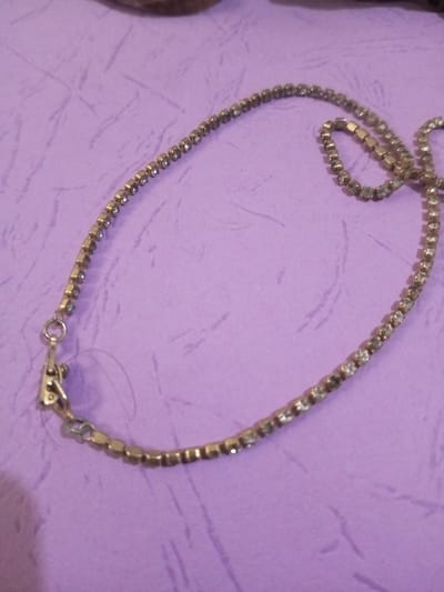 The image displays a single metallic chain-style necklace or bracelet, characterized by a continuous line of small, clear, faceted stones, possibly rhinestones, set in dull gold- or brass-toned metal. The item is laid out in an irregular, elongated curve on a light purple, textured fabric or paper surface. A simple hook-and-loop clasp, consistent in color with the rest of the metal, is visible on the left end of the chain. A thin, light-colored filament is present near the clasp. In the upper portion of the frame, an out-of-focus, brownish, textured object is partially visible. The scene is located in Chandragiri, Nepal.